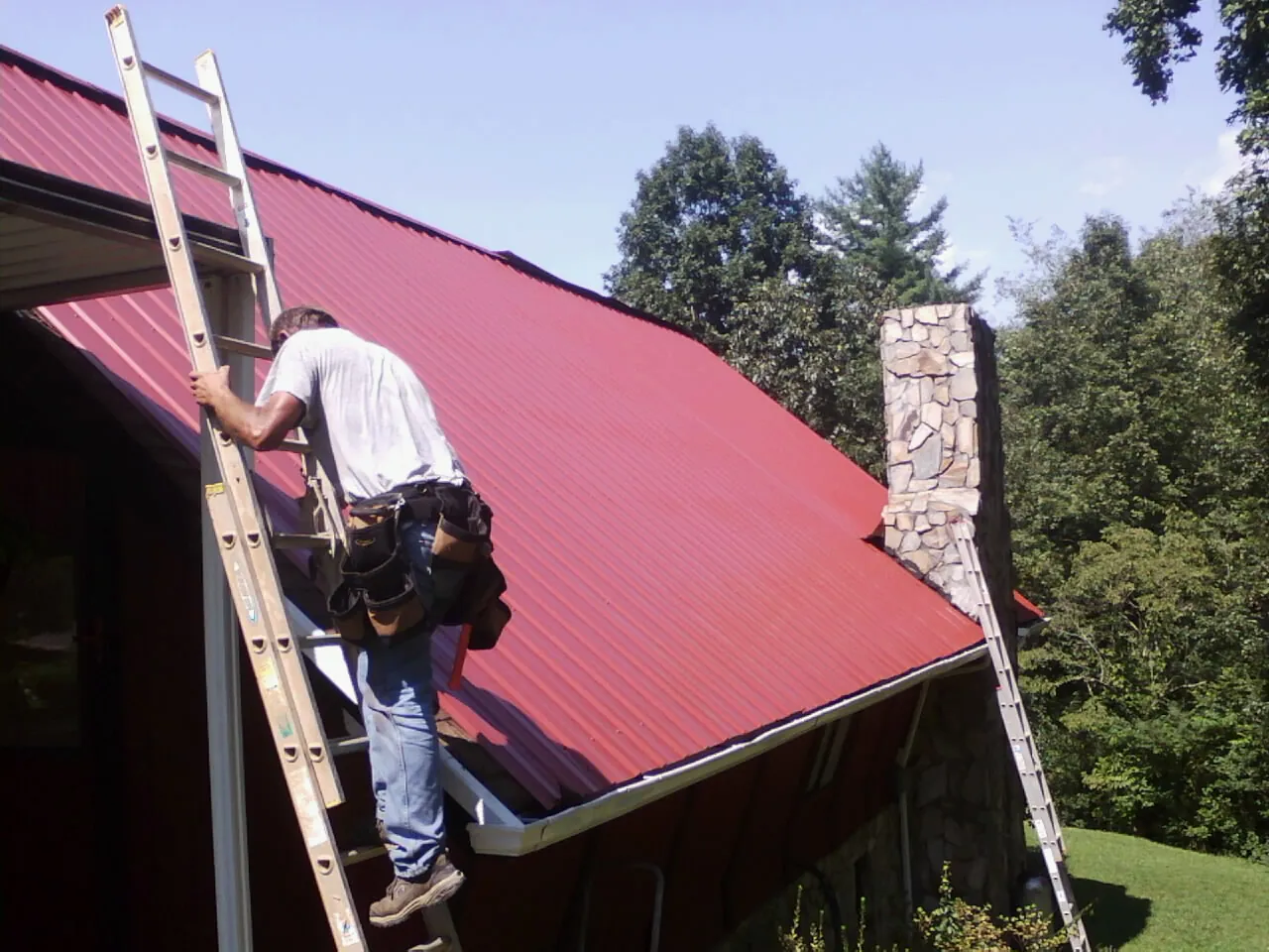 Roofer on a ladder beside a red metal roof and stone chimney — Soffit & Fascia Repair by Roofing Brothers in Barnard